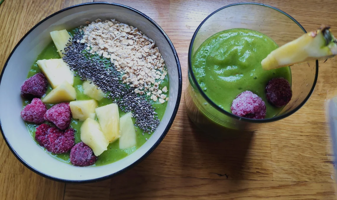 Eine Smoothie Bowl und ein Glas mit grünem Smoothie stehen auf einem Holztisch.